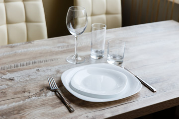 Table setting. Two white plates, one on top of another. Knife and fork. Wine glass, water glass and old fashioned glass. Wooden table. Selective focus.