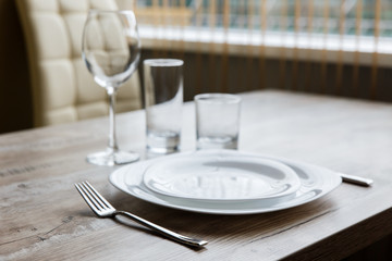 Table setting. Two white plates, one on top of another. Knife and fork. Wine glass, water glass and old fashioned glass. Wooden table. Selective focus.