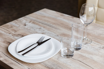 Table setting. Two white plates, one on top of another. Knife and fork. Wine glass, water glass and old fashioned glass. Wooden table. Selective focus.