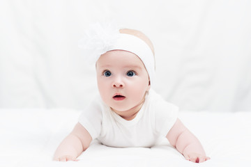 Cute baby girl in white shirt, with bow flower on head, lying on belly on soft blanket, indoors