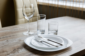 Table setting. Two white plates, one on top of another. Knife and fork. Wine glass, water glass and old fashioned glass. Wooden table. Selective focus.