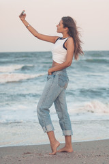 Young girl with beautiful figure is traveling and making selfies on the beach at the sunrise. Beautiful sky background.