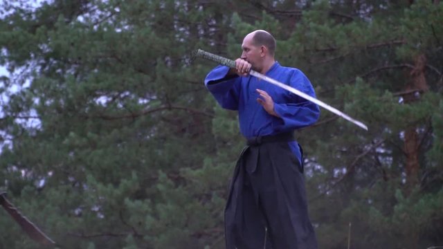 A confident bald samurai in with a blue kimono walks through the forest and becomes in the fighting position that would fight against a warrior with a halberd
