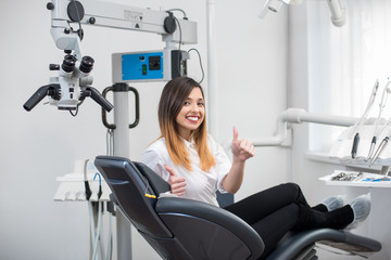 Naklejka premium Happy female patient with perfect white teeth sitting in dental chair, smiling and showing thumbs up after treatment at modern dental clinic. Dentistry, medicine and healthcare concept.