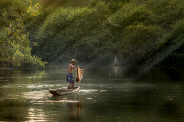 Thai fisherman on wooden boat casting a net for catching freshwater fish in nature river in the early evening before sunset