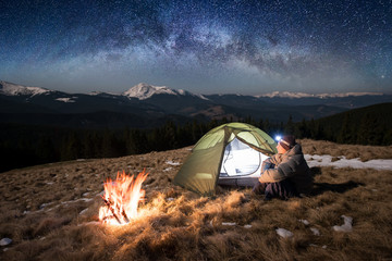 Fototapeta Male tourist enjoying in his camp at night. Man with a headlamp sitting near campfire and tent under beautiful sky full of stars and milky way. On the background snow-covered mountains obraz