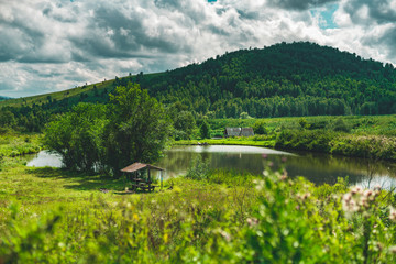 Obraz premium Summer landscape with small lake of fresh water, hills with forests in distance, green meadows of native grasses, arbour with trees near, submerged country house, Altai mountains, Aya district, Russia