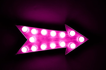 Pink vintage bright and colorful illuminated display arrow sign with light bulbs against a pink dark background