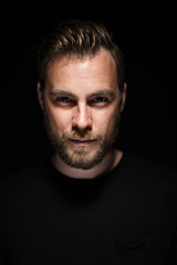 Portrait of a bearded man with an intense look standing in a dark room against a black background.