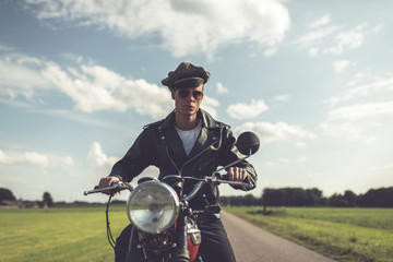 Vintage motorcyclist in sunglasses and cap riding in countryside.