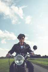 Vintage motorcyclist in sunglasses and cap riding in countryside.
