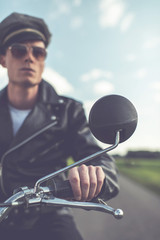 Vintage motorcyclist in sunglasses and cap riding in countryside.