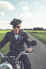 Vintage motorcyclist in sunglasses and cap riding in countryside.