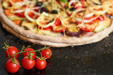 Hot vegetarian pizza with tomatoes, bell pepper, onion, black olives, cheese, spices on black dark blurred background close up decorated with fresh basil and cherry tomatoes on foreground. Side view