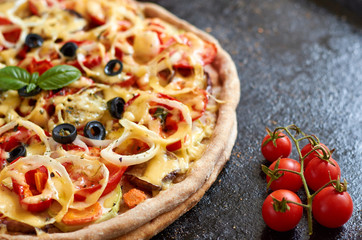 Hot vegetarian pizza with tomatoes, bell pepper, onion, black olives, cheese and spices on black dark background close up decorated with fresh basil and cherry tomatoes. Side view