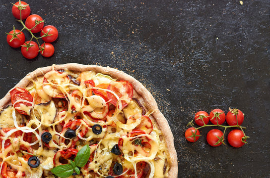 Hot vegetarian pizza with tomatoes, bell pepper, onion, black olives, cheese, spices on dark black baking tray background decorated with small cherry tomatoes close up with copy space. Top view