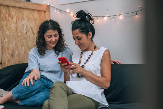 Two women friends use a smartphone in apartment on sofa