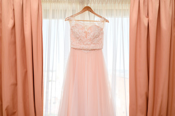 Beautiful lace wedding dress hanging on hanger on window with curtains in room, free space