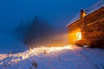 Wooden Koliba house with ray of warm light from the window in evening snowy winter forest of Carpathians mountains, Ukraine