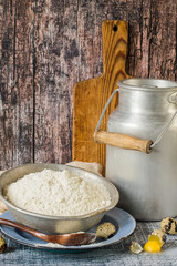  Aluminum jar with cow's milk, a bowl of flour, quail eggs, a plate and a wooden cutting board on an old table.