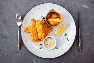 Fish and chips on a white plate with fork and knife on dark background