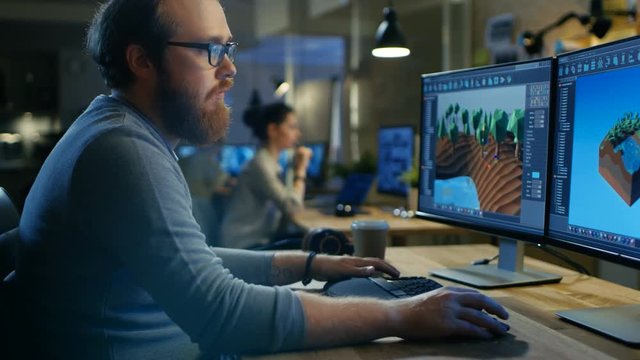 Creative Male Game Developer Works with Graphics for the New Level Design, He Sits at His Wooden Table Working on Two Display Personal Computer. Other People Work in This Creative Studio.