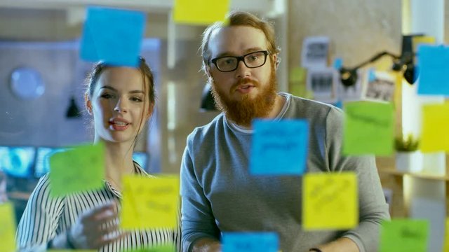 Male and Female Designers Discuss and Outline They're Project with Sticky Notes on a Glass Wall. They're Young and Creative People Who Work in a Stylish Loft Space.