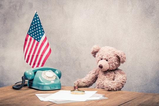 Retro Teddy Bear toy sitting at the desk with telephone, paper blanks, magnifying glass and USA flag front concrete wall background. Vintage instagram style filtered photo