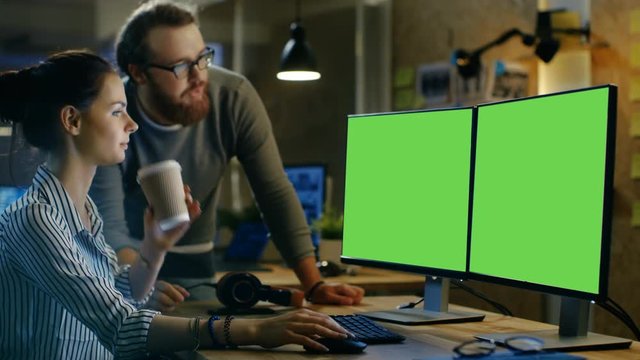Beautiful Young Woman Works on a Isolated Mock-up Green Screen Personal Computer, Her Bearded Male Colleague Supports Her and Makes Conversation. They Work in a Creative Loft Office.