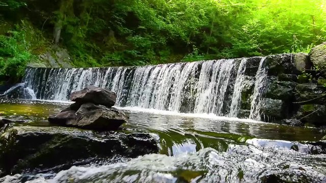 Beautiful small waterfall in forest stream in South Czech. Green nature, summertime. Sun lights on water surface and in trees. Rocks in water, nice waves.