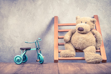 Retro Teddy Bear standing on wooden ladder and toy bicycle in front concrete wall background. Vintage instagram style filtered photo