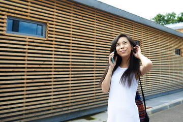 Outdoor picture of beautiful female student of Asian appearance walking by wooden building construction, speaking on mobile phone with her friend while spending summer vacations in resort town alone