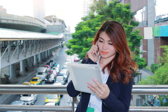 Attractive Young Asian Business Woman Talking On Phone And Looking Document Files In Her Hands At Outside Office Background.