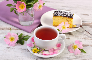 Cup of tea with cheesecake and wild rose flower on old board