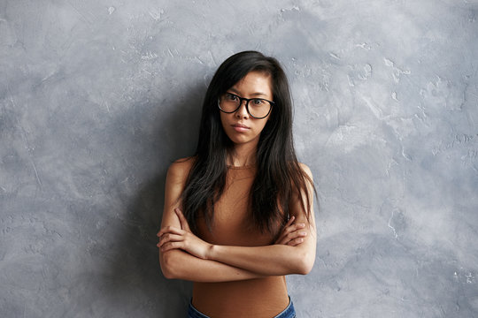 Portrait of offended stubborn young brunette Asian woman wearing eyeglasses standing isolated at grey textured wall, keeping arms folded, looking at camera with angry expression on her face
