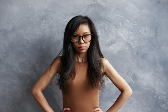 Studio shot of annoyed irritated brunette girl of Asian appearance wearing eyeglasses posing at grey wall with hands on her waist, feeling mad and furious about something, hel look full of anger