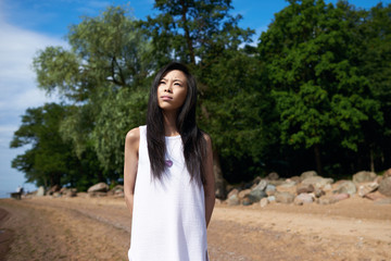 Gorgeous young Japanese woman spending holidays by the ocean, walking along shore, having thoughtful serious look, breathing fresh sea air. Pensive Chinese student girl enjoying summer vacations