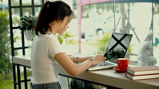 Side view of young pretty girl is working or chatting on a laptop by cafe window while sitting and drinking hot coffee