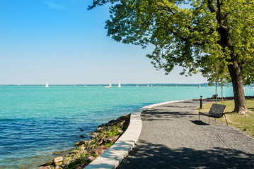 A view of Balaton lake with white yachts at the horizon and trees, bench and footpath at the foreground, Tihany, Hungary.