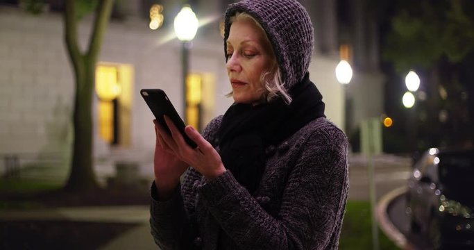 Attractive senior woman wrapped in scarf and sweater messaging on cellphone outdoors in evening. Happy elderly woman texting someone outside building on quiet night. 4k 