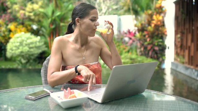 Attractive woman looks absorbed while using notebook and drinking orange juice, steadycam shot

