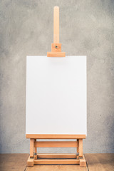 Fototapeta premium Portable desk easel for painting with canvas blank on wooden table front concrete wall background. Retro instagram old style filtered photo
