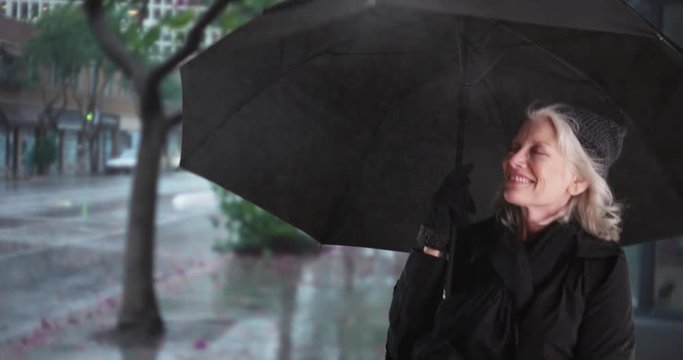 Carefree senior woman with umbrella dancing and jumping outside in the rain smiling. Joyful elder woman having fun outdoors on rainy day spinning with umbrella on wet sidewalk. 4k 