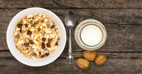 White bowl with Granola or Muesli and a jar of milk or plain yogurt with a spoon on a vintage wood background. Healthy breakfast top view composition with copy space.