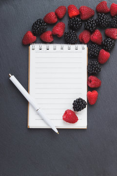 Red raspberries, blackberry, notebook and a pen on the black slate stone background