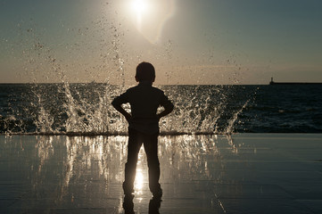 Silhouette of a small boy against the backdrop of a huge wave lit by the sunset