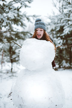 young teenage hipster girl having fun, make snowman in winter time forest, wearing sweaters and scarfs, outdoors, snowy weather