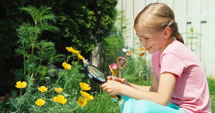 Cheerful girl 8 aged looking at yellow flower through magnifying glass outdoors. Child drinking juice from glass
