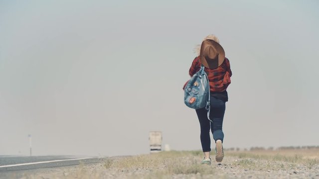 Young stylish girl in cowboy clothes doing hitch-hiking in desert, on the road. She carries backpack and hat. Pass trucks, delivery trucks. California or Nevada. Search yourself, escape from problems