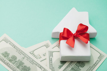 Small white house decorated red bow ribbon and dollars money on green background. Buying a new home, gift or sale of real estate concept.